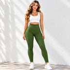 A woman modeling high-waisted ribbed green leggings with a white sports bra and white sneakers, standing against a white textured wall with soft leaf shadows.