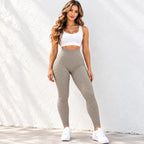 A woman modeling a matching activewear set consisting of a white sports bra and textured high-waisted leggings in a warm taupe-beige color, paired with white sneakers, standing against a white textured wall.