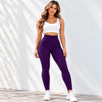 A woman modeling a matching activewear set consisting of a white sports bra and high-waisted purple textured leggings, standing against a white textured wall with leaf shadows.