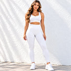 A woman modeling a white activewear set consisting of a sports bra and textured leggings, paired with white sneakers, standing against a white textured wall.