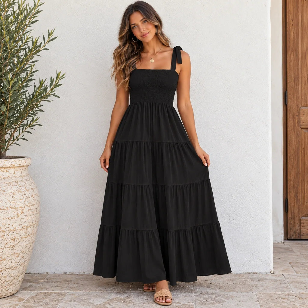 A woman wearing a black tiered maxi dress with a square neckline and shoulder tie details, standing against a white stucco wall next to a potted plant.