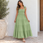 A woman wearing a light sage green tiered maxi dress with tie-shoulder straps, standing outdoors against a white stucco wall next to a large potted plant.