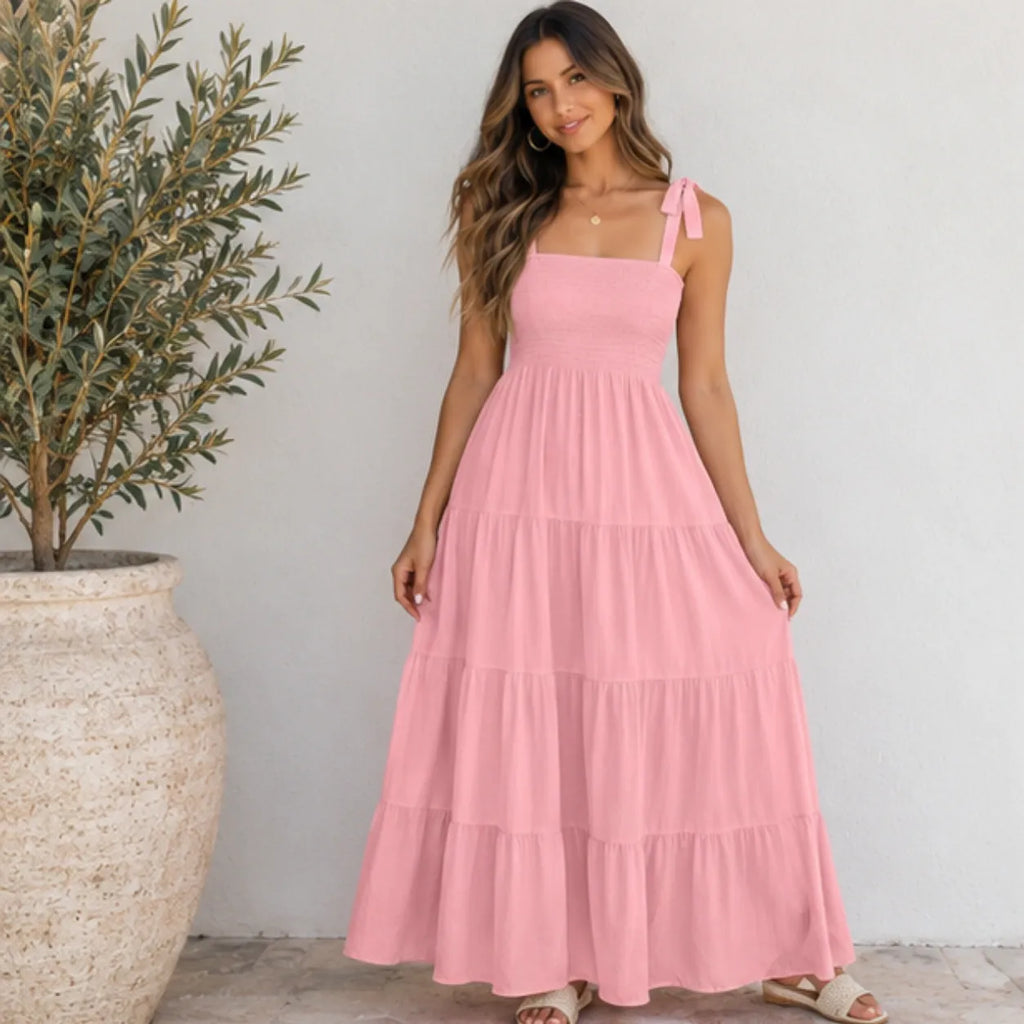 A woman wearing a light pink tiered maxi dress with tie-shoulder straps, standing in front of a white wall next to a large potted olive tree.