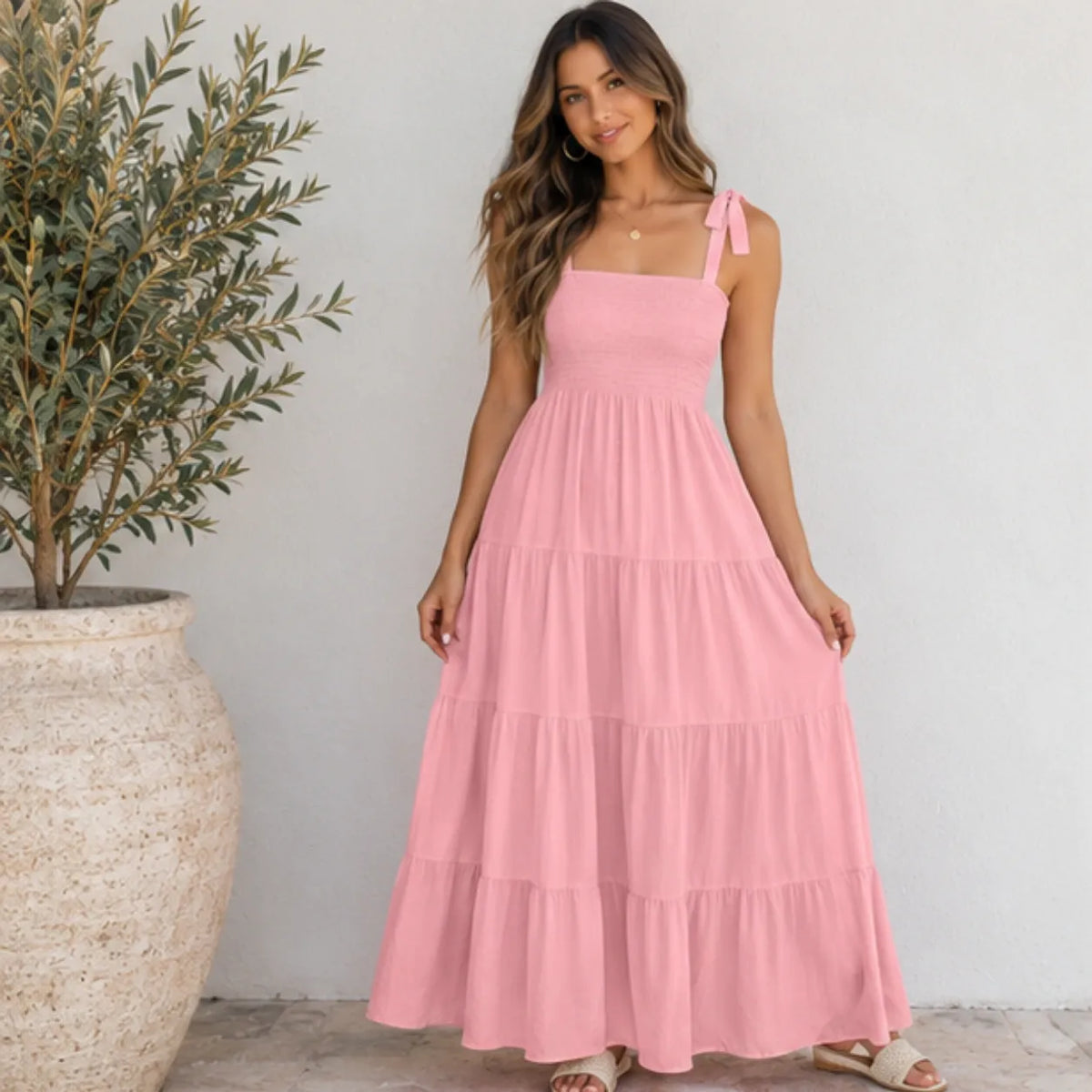 A woman wearing a light pink tiered maxi dress with tie-shoulder straps, standing in front of a white wall next to a large potted olive tree.