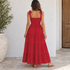 A woman viewed from behind wearing a red sleeveless maxi dress with tie shoulder straps and a tiered skirt, standing on a stone patio against a white wall.