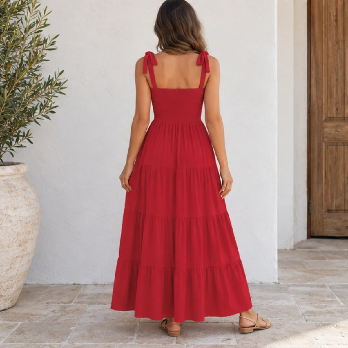 A woman viewed from behind wearing a red sleeveless maxi dress with tie shoulder straps and a tiered skirt, standing on a stone patio against a white wall.