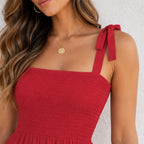 A close-up of a woman wearing a red smocked sleeveless top or dress with tie-shoulder straps, paired with a gold pendant necklace, against a light textured wall.
