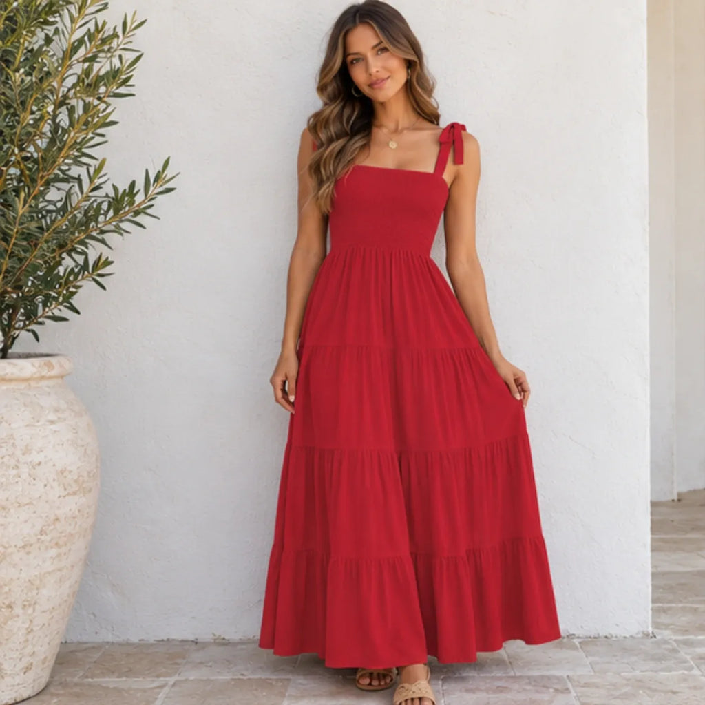 A woman wearing a red tiered maxi dress with tie-shoulder straps, standing against a white stucco wall next to a large potted olive tree.