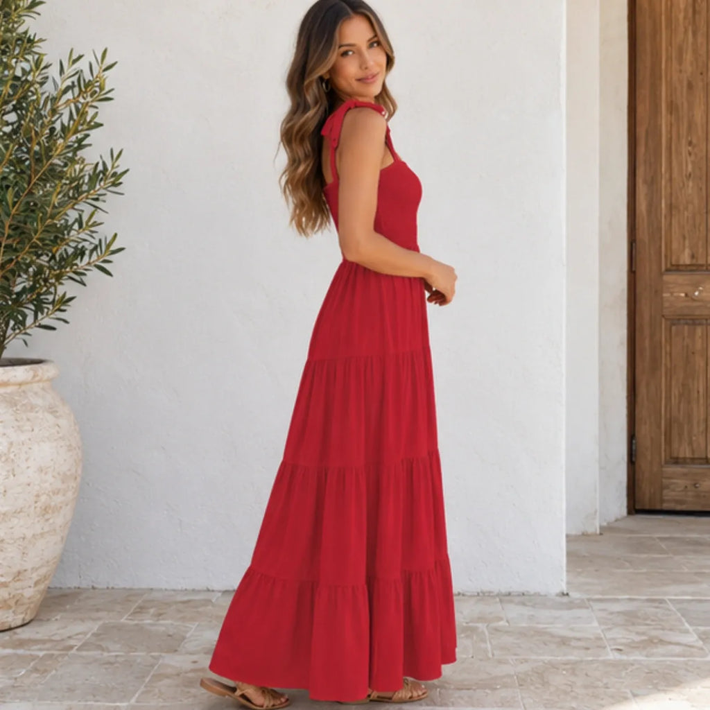 A woman wearing a sleeveless, tiered red maxi dress stands in a sunlit outdoor setting with a white wall, tiled floor, and potted plant in the background.