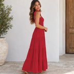 A woman wearing a sleeveless, tiered red maxi dress stands in a sunlit outdoor setting with a white wall, tiled floor, and potted plant in the background.