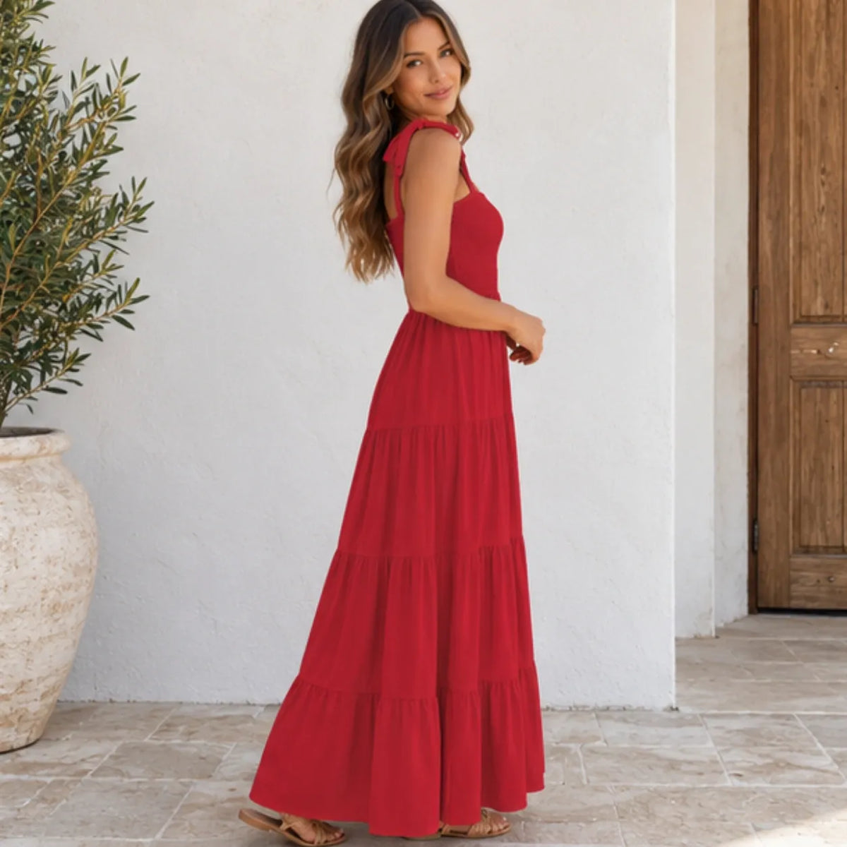 A woman wearing a sleeveless, tiered red maxi dress stands in a sunlit outdoor setting with a white wall, tiled floor, and potted plant in the background.
