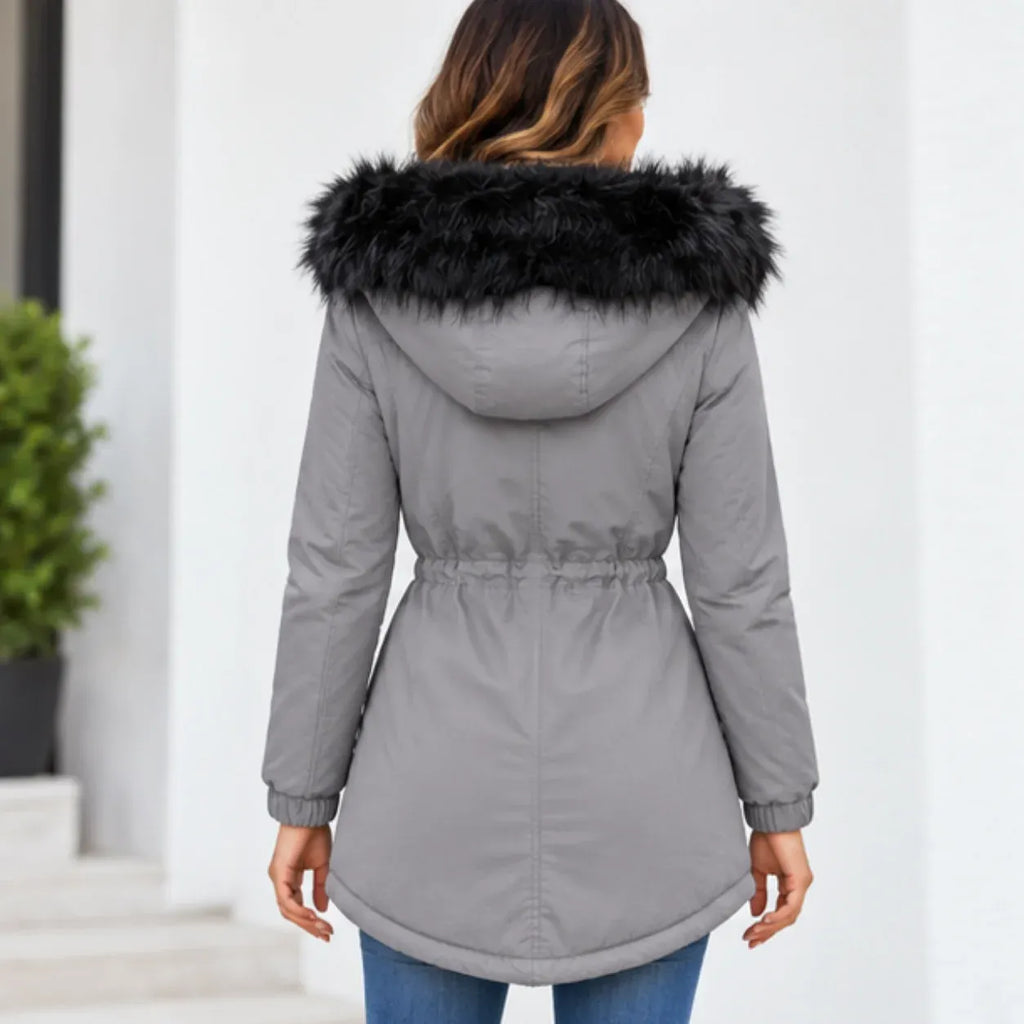A woman viewed from behind wearing a gray hooded puffer coat with a black faux-fur trim, paired with blue jeans, set against a bright white building background.