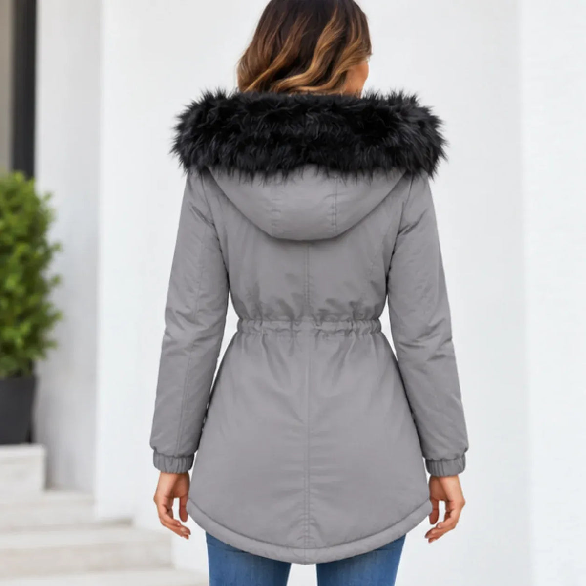 A woman viewed from behind wearing a gray hooded puffer coat with a black faux-fur trim, paired with blue jeans, set against a bright white building background.