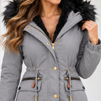 A close-up of a woman wearing a gray hooded winter parka with a black faux-fur trimmed hood, black faux-fur lining, gold-tone snap buttons, and drawstring details.