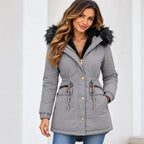 A woman wearing a gray hooded puffer parka with a black faux-fur trimmed hood, black lining, gold-tone snap buttons, and drawstring details, paired with blue jeans against a light gray background.