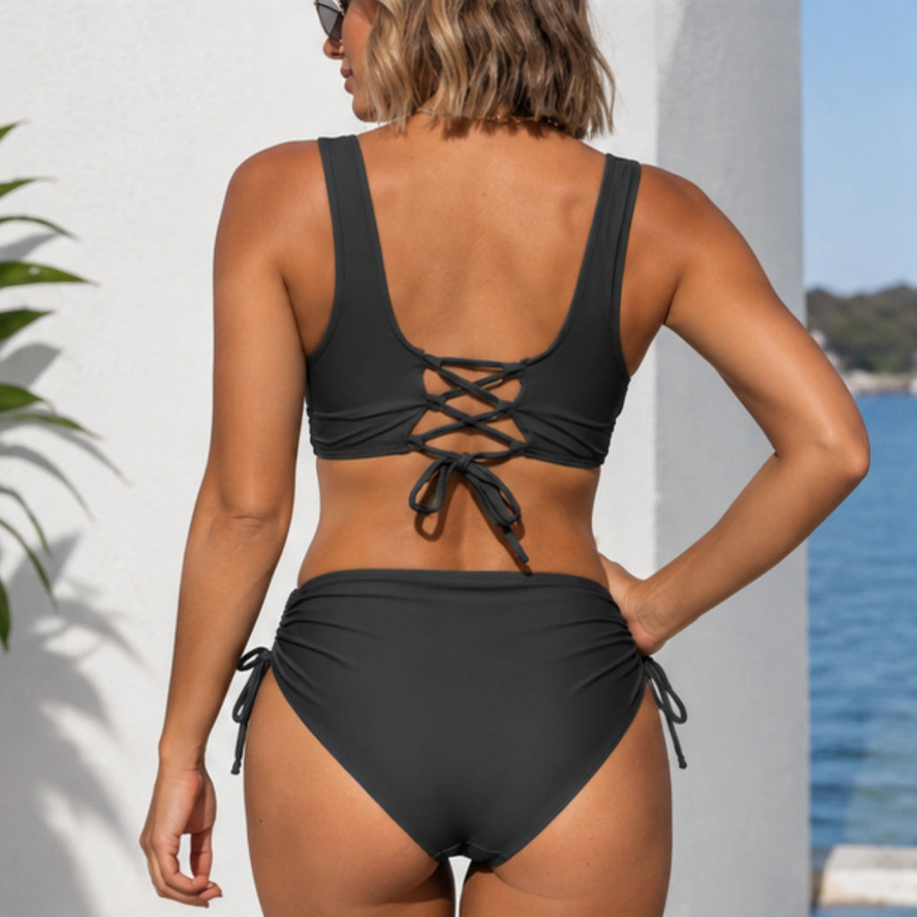 A woman shown from the back wearing a black two-piece bikini with lace-up back detail and side-tie bottoms, standing outdoors near a white wall and blue water.