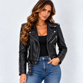A woman modeling a black faux leather moto jacket with silver zippers and snap details, paired with a black top and blue jeans, against a light gray studio background.