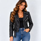 A woman modeling a black faux leather moto jacket with silver zippers and snap details, paired with a black top and blue jeans, against a light gray studio background.