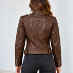 A woman viewed from the back wearing a brown faux-leather moto jacket with silver zipper details and dark gray jeans, set against a plain light gray background.