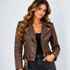 A woman wearing a brown faux leather moto jacket with silver zippers and snap details, styled over a black top and dark jeans, against a light gray studio background.