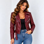 A woman modeling a cropped, asymmetrical moto jacket in burgundy faux leather, styled with a black top and blue jeans against a plain white studio background.