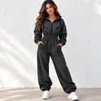 A woman modeling a black zip-up hooded sweatshirt and matching wide-leg sweatpants set, standing against a white textured wall.
