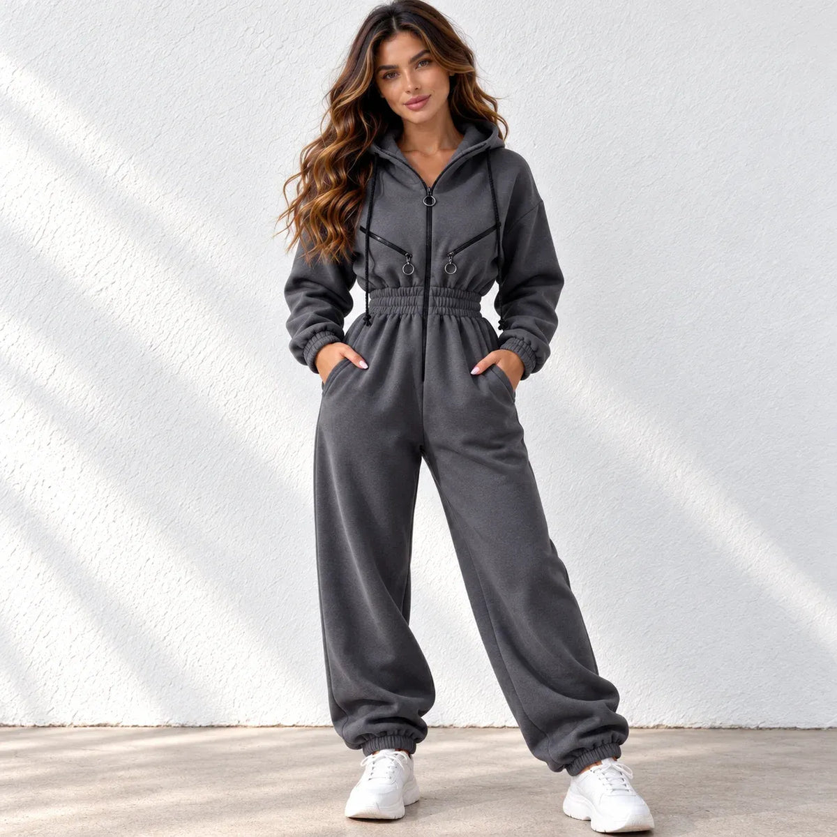 A woman modeling a dark gray zip-up hooded sweatshirt and matching jogger pants set, standing against a white textured wall with soft diagonal shadows.