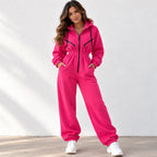 A woman modeling a bright pink zip-up hooded sweatshirt and matching jogger pants set, styled with white sneakers against a white textured wall.