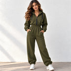A woman modeling an olive green zip-up hooded sweatshirt and matching jogger pants set, standing against a textured white wall.