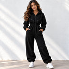 A woman modeling a black zip-up hoodie sweatshirt with matching wide-leg sweatpants, styled with white sneakers against a white textured wall with soft shadows.