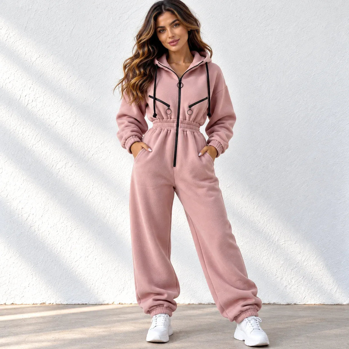 A woman modeling a dusty pink fleece tracksuit with black zipper and drawstring accents, paired with white sneakers, standing against a white textured wall.