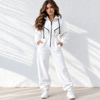 A woman modeling a white zip-up hoodie and matching sweatpants set with black drawstrings and zipper accents, standing against a white textured wall with soft shadow patterns.