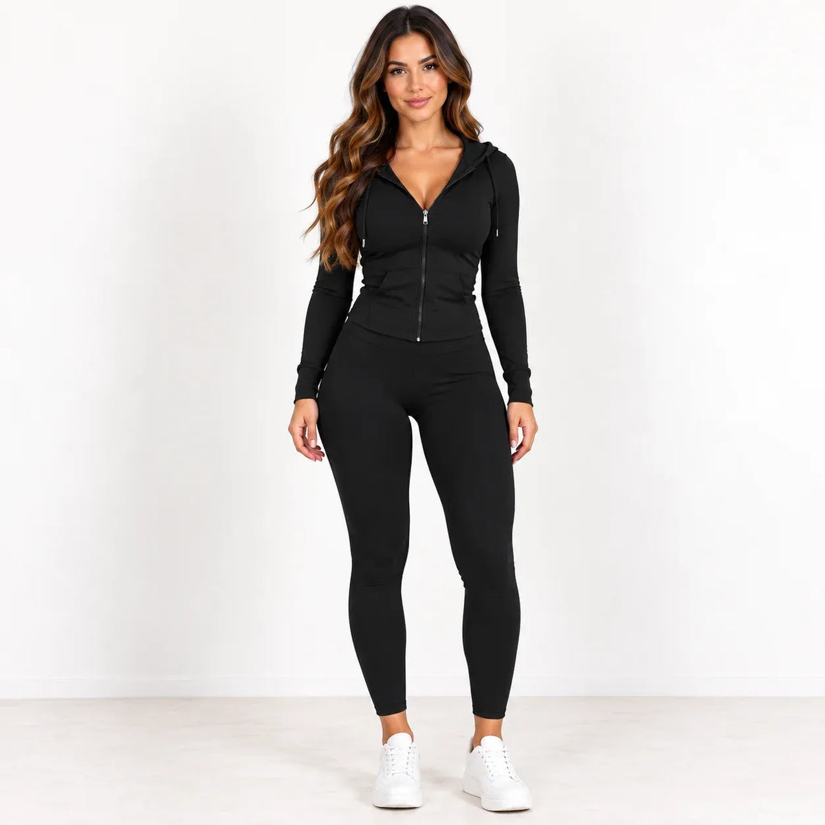 A woman modeling a black long-sleeve zip-up hoodie and matching black leggings with white sneakers, standing against a plain white studio background.