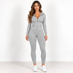 A woman modeling a light gray two-piece activewear set consisting of a zip-up long-sleeve hoodie and matching leggings, paired with white sneakers, against a plain white studio background.