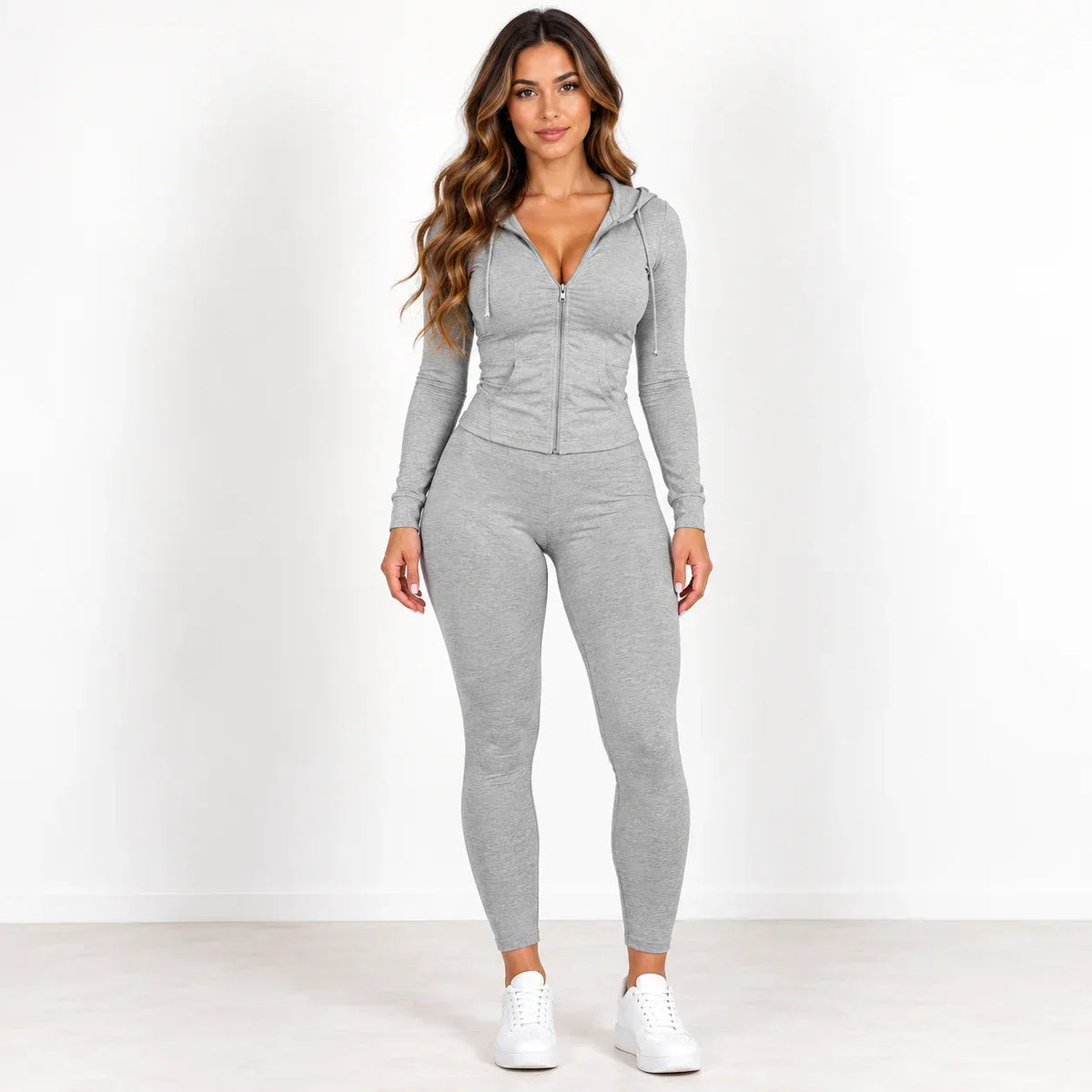 A woman modeling a light gray two-piece activewear set consisting of a zip-up long-sleeve hoodie and matching leggings, paired with white sneakers, against a plain white studio background.