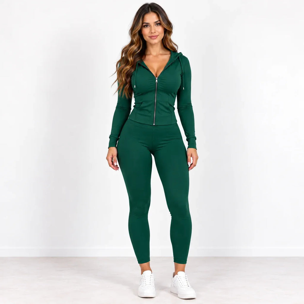 A woman modeling a dark green two-piece activewear set consisting of a zip-up long-sleeve hoodie and matching high-waisted leggings, paired with white sneakers, against a plain white studio background.