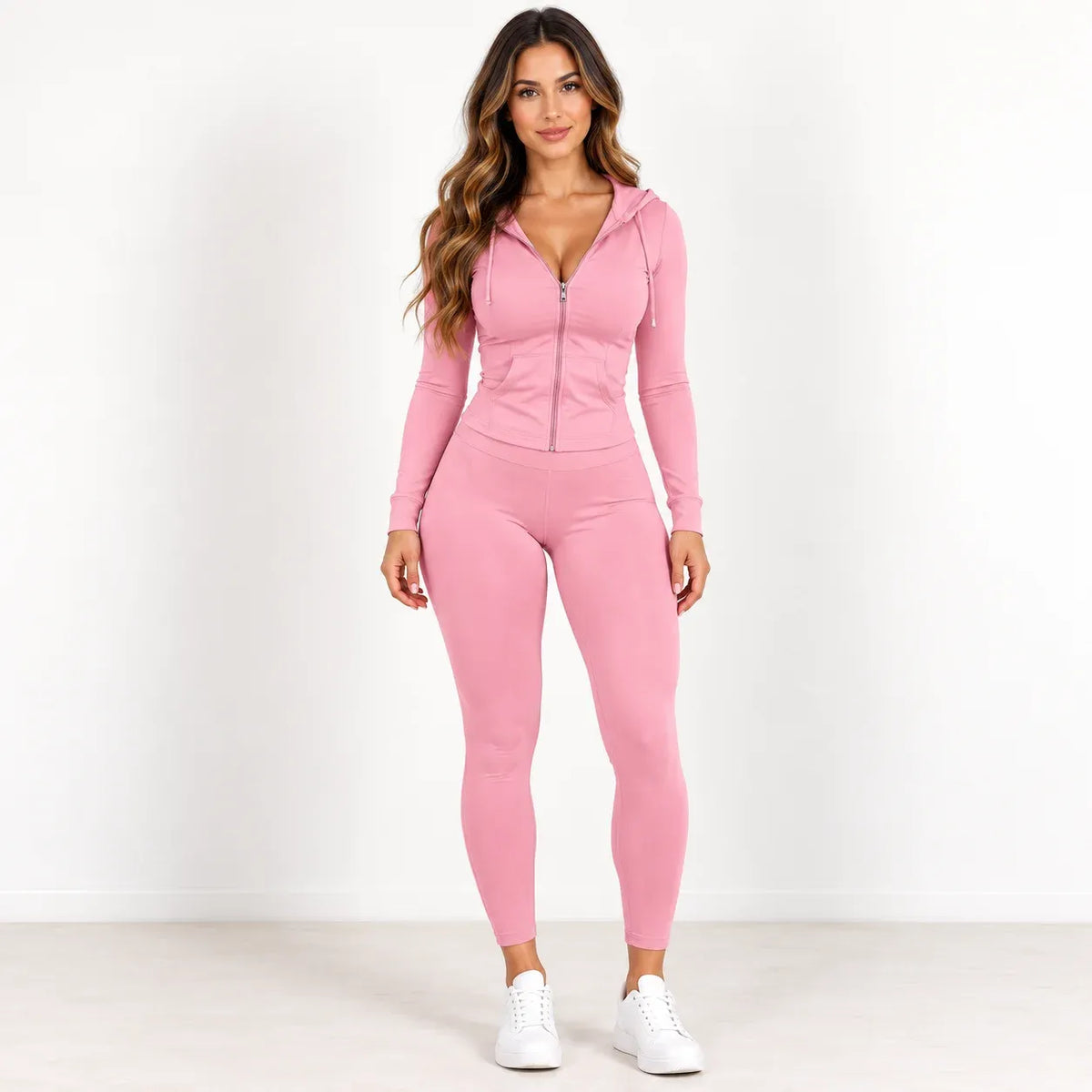 A woman modeling a matching pink long-sleeve zip-up hoodie and high-waisted leggings set, paired with white sneakers, against a plain white studio background.