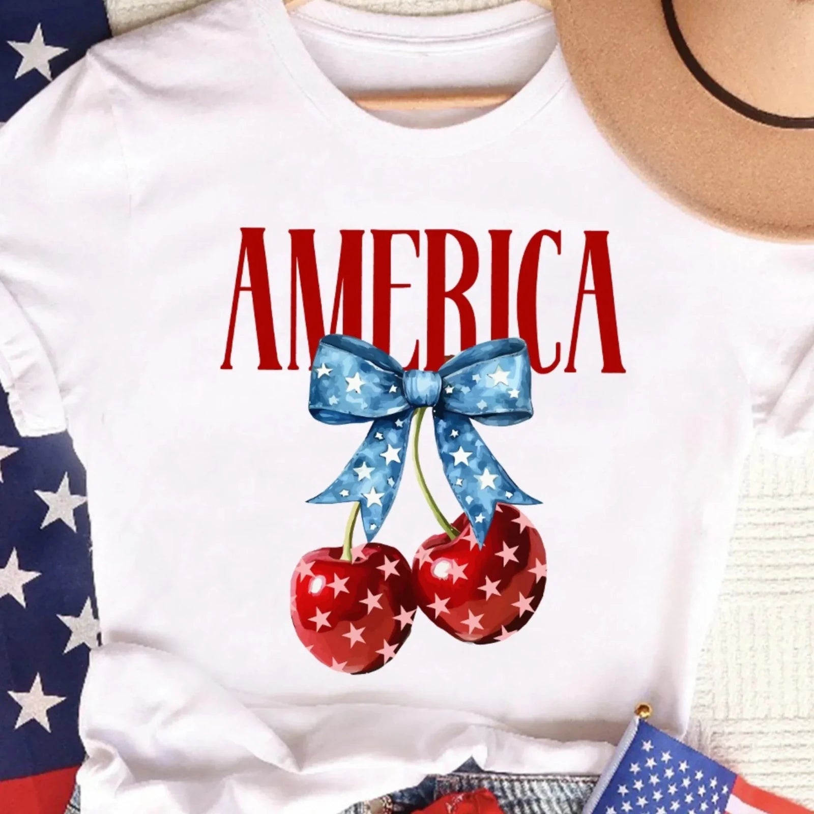 Flat lay of a white women's America graphic T-shirt featuring patriotic cherries with a blue star bow, styled with American flag accessories and denim shorts.
