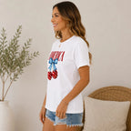 Side view of a women's white patriotic America graphic T-shirt with star-spangled cherries and blue bow, paired with distressed denim shorts.