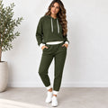 A woman modeling a matching olive green waffle-knit lounge set, consisting of a cropped hoodie with white drawstrings and white ribbed trim, paired with matching jogger pants, white sneakers, and standing in a bright room next to a potted plant.