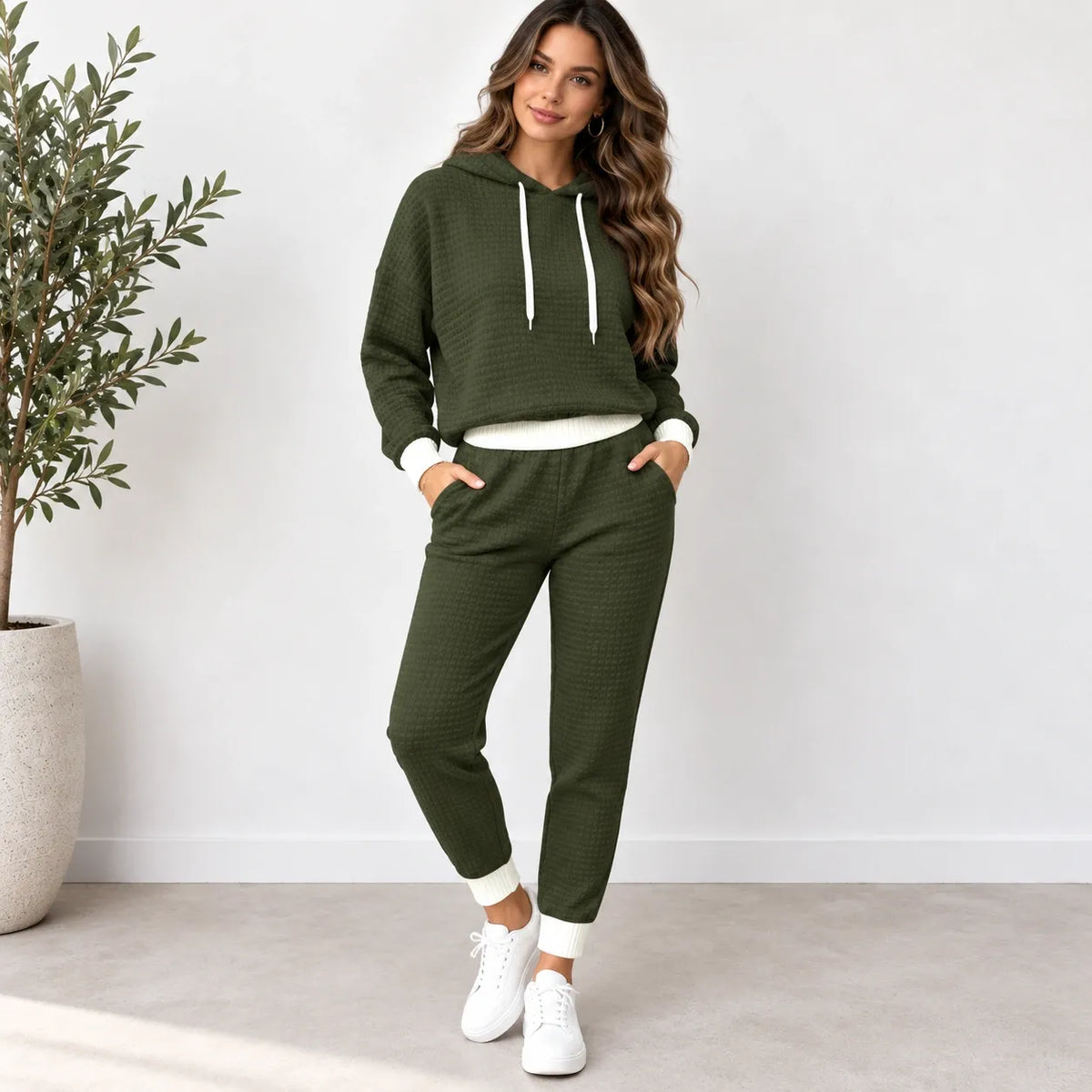 A woman modeling a matching olive green waffle-knit lounge set, consisting of a cropped hoodie with white drawstrings and white ribbed trim, paired with matching jogger pants, white sneakers, and standing in a bright room next to a potted plant.