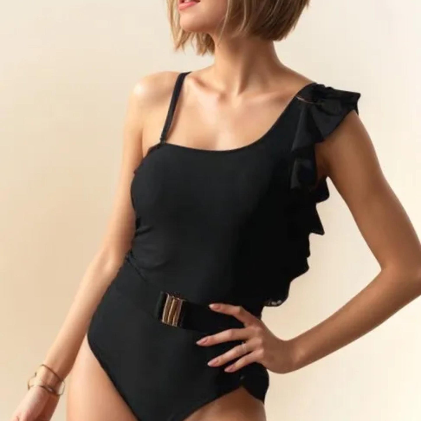 Lifestyle photo of a woman wearing a black asymmetric one-shoulder swimsuit on a rocky beach.