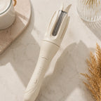 Ivory automatic rotating curling iron displayed on a marble vanity with beauty accessories.