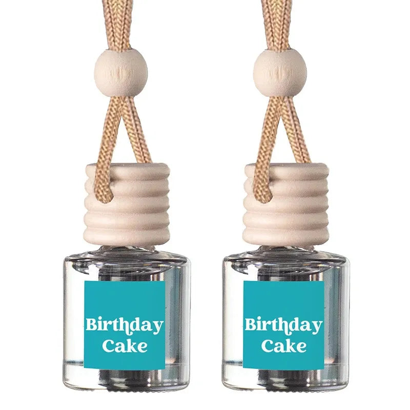 Two clear glass reed diffuser bottles with beige wooden caps, hanging cords, and turquoise labels reading 'Birthday Cake' on a white background.