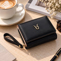 A black faux-leather wallet with a wrist strap and gold-tone hardware, styled on a beige tabletop with a coffee cup, sunglasses, and dried flowers in the background.