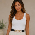 Woman wearing a black elastic waist belt with a textured gold chain buckle, styled with a white ribbed tank top and cream trousers, front view.