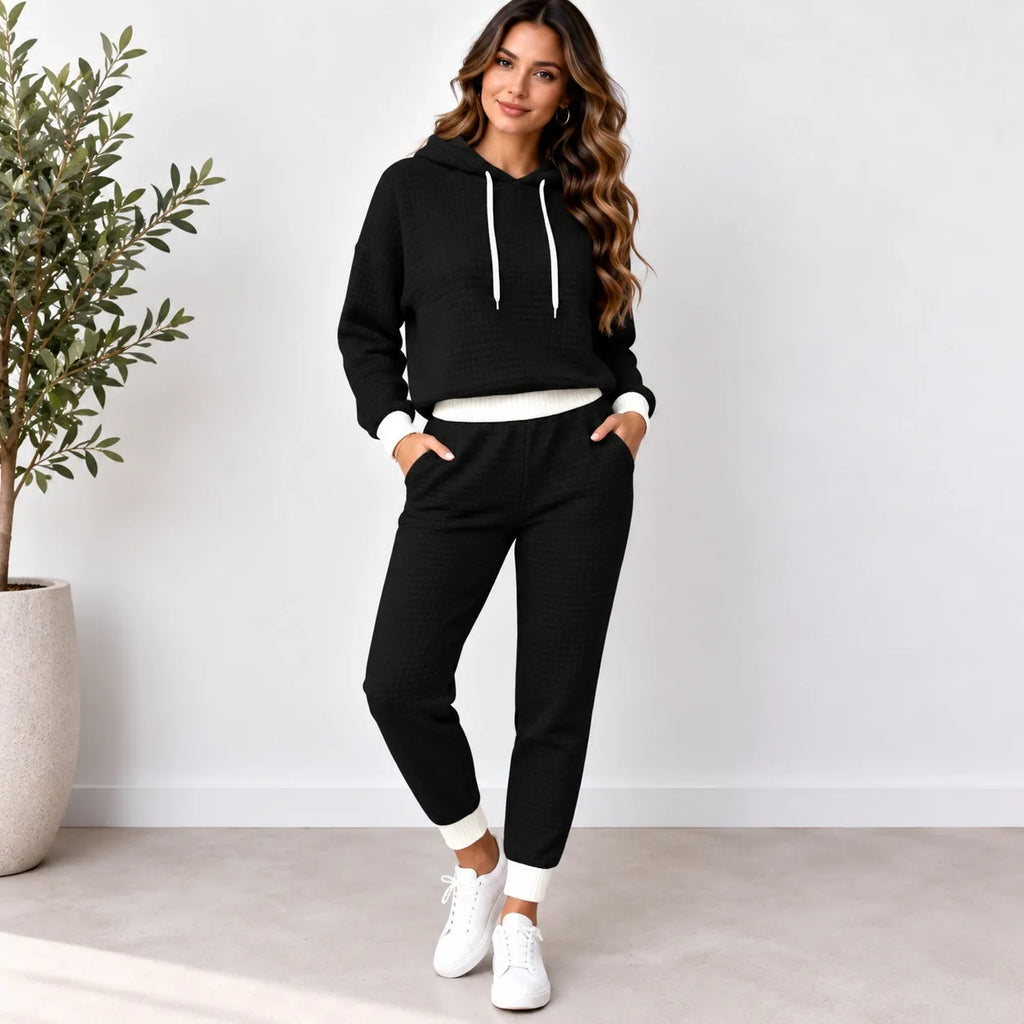 A woman modeling a black ribbed two-piece loungewear set consisting of a cropped pullover hoodie with white drawstrings and matching jogger pants with white cuffs, standing in a bright room with a potted plant.