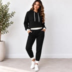 A woman modeling a black ribbed two-piece loungewear set consisting of a cropped pullover hoodie with white drawstrings and matching jogger pants with white cuffs, standing in a bright room with a potted plant.