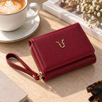 A deep red faux-leather wallet with a wrist strap and gold deer logo, styled on a beige tabletop next to a coffee cup, book, and dried flowers.
