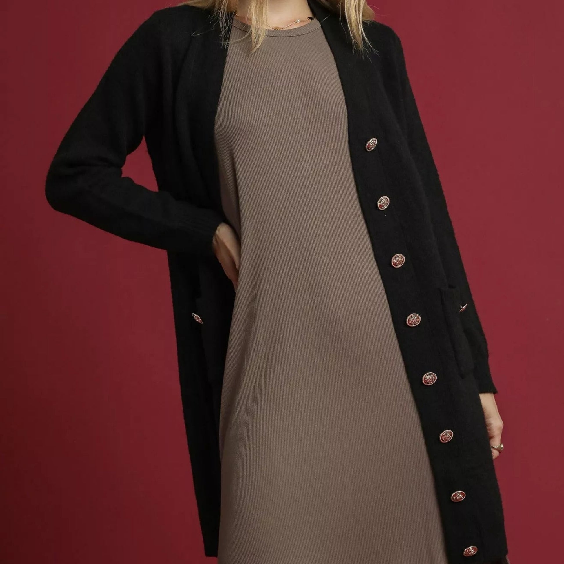 Person wearing a black coat with gold buttons over a brown dress on a red background